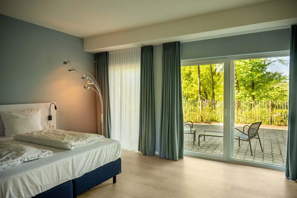 Modernes Hotelzimmer mit einem Doppelbett mit weißen Bettwäsche, einer mehrarmigen Stehlampe neben dem Bett, grauen Vorhängen und großen Fenstern, die auf eine Terrasse mit Stühlen und grünem, sonnigem Gartenbereich hinausblicken.