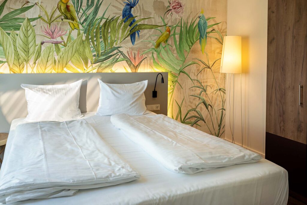 Hell eingerichtetes Schlafzimmer mit einem Doppelbett, weißen Bettdecken und Kissen, einer Wand mit farbenfrohem tropischem Wandbild und einer Stehlampe in der Ecke neben einem Holzkleiderschrank.