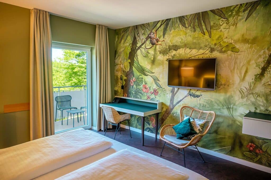 Ein Hotelzimmer mit grüner Wandmalerei einer Dschungelszene, einem Fernseher an der Wand, einem Schreibtisch, einem Rattan-Stuhl, einem gepolsterten Stuhl mit Kissen, einem Bett, und einem Balkon mit Blick auf grüne Bäume.