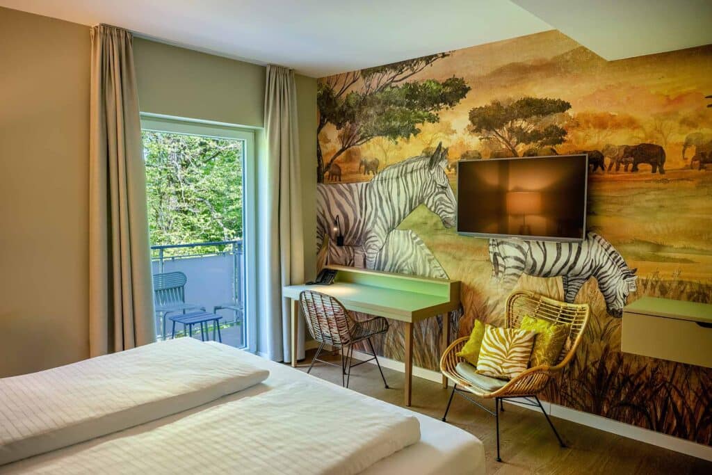 Zimmer mit tropischer Wildtier-Wandmalerei, schwarzem Flachbildfernseher, Schreibtisch mit Telefon, Sessel mit Kissen und Balkon mit Blick auf grüne Bäume durch offene Türen.