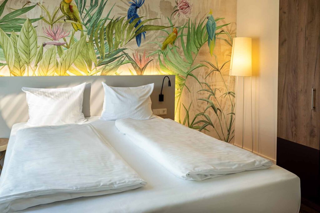 Ein Hotelzimmer mit einem weißen Bett, einem Kopfteil mit einer Wanddekoration aus einer tropischen Pflanzentapete, einem Wandlampenarmatur und einem Stehlampe neben einem Holzschrank.