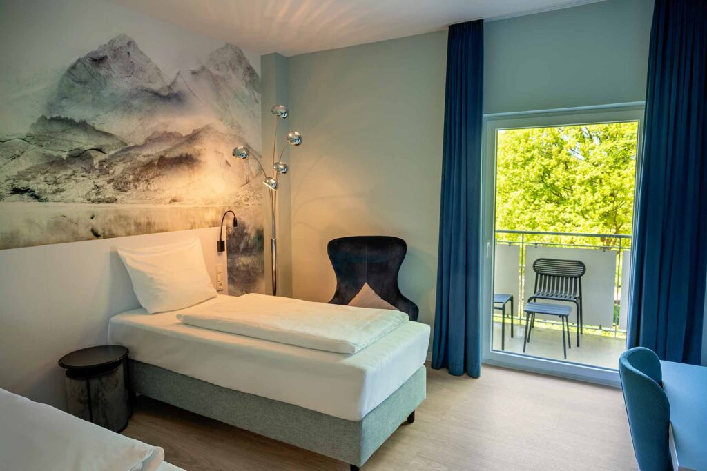 Ein modernes Hotelzimmer mit zwei Einzelbetten, einem Wandbild von Bergen, einem schwarzen Sessel, einem Nachttisch, einer Stehleuchte und einem Balkon mit zwei Stühlen, durch den man auf grüne Bäume draußen sieht.