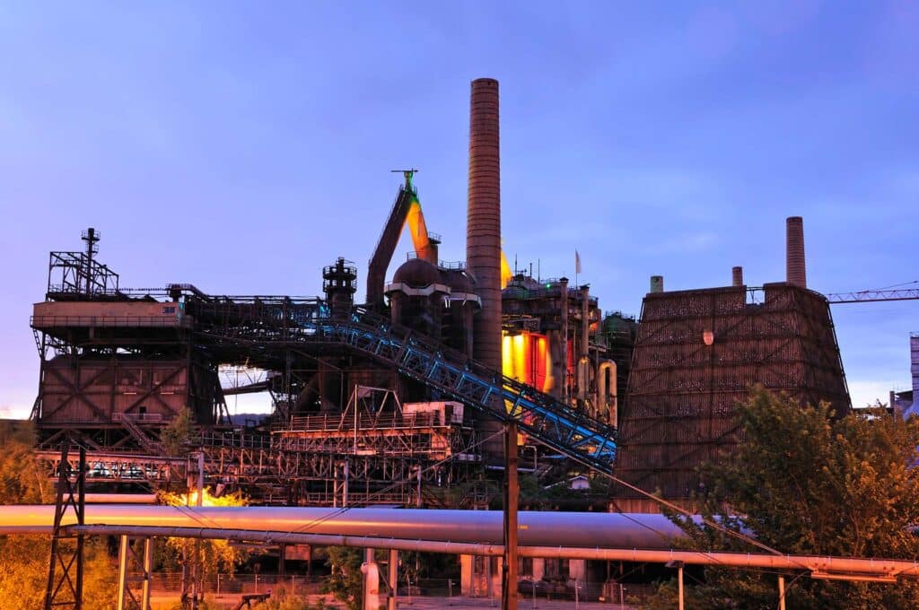 Industrielle Fabrikanlage bei Sonnenuntergang mit leuchtenden orangefarbenen und blauen Lichtern, viele Schornsteine und komplexe Metallstrukturen.